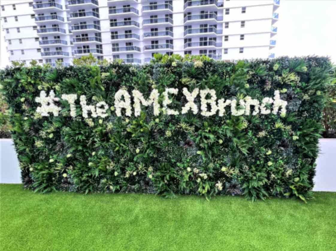 A bespoke faux green wall adorned with white flowers spelling #TheAMEXBrunch graces a lush lawn in Miami. Tall buildings rise majestically in the background, enhancing the scenic Florida setting.