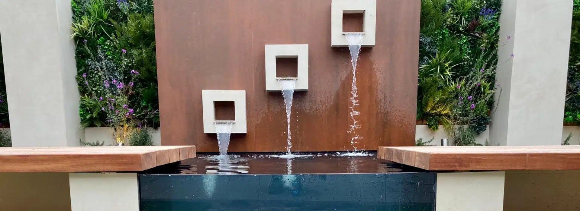 A modern water feature with three square spouts cascades water into a rectangular pool, set against a rust-colored wall framed by lush plants and purple flowers. Pale pillars border each side, enhancing the contemporary design of this private gardens green wall in Cheshire.