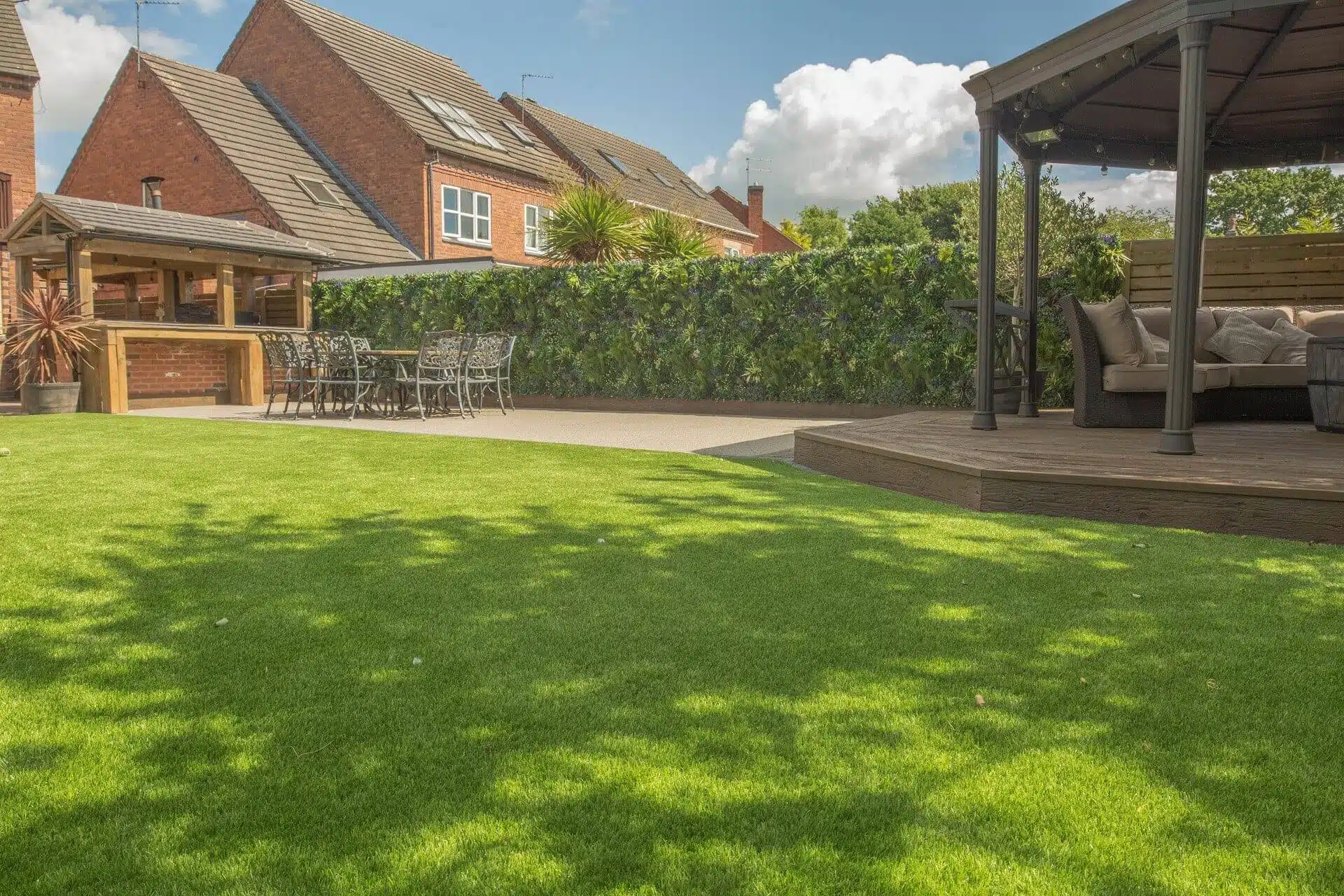 A backyard with a lush green lawn, a wooden gazebo with a sofa on the right, and a patio table set under an awning on the left. Bordered by plants, shrubs, and a garden fence, brick houses are visible in the background under Staffordshires blue sky with clouds.