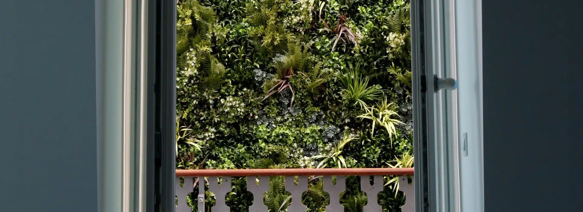 Through the window, a lush green wall serves as privacy screening in Portugal, showcasing a vertical garden filled with diverse plants. In the foreground, a decorative balcony railing with geometric cutouts adds charm to this serene and natural scene.