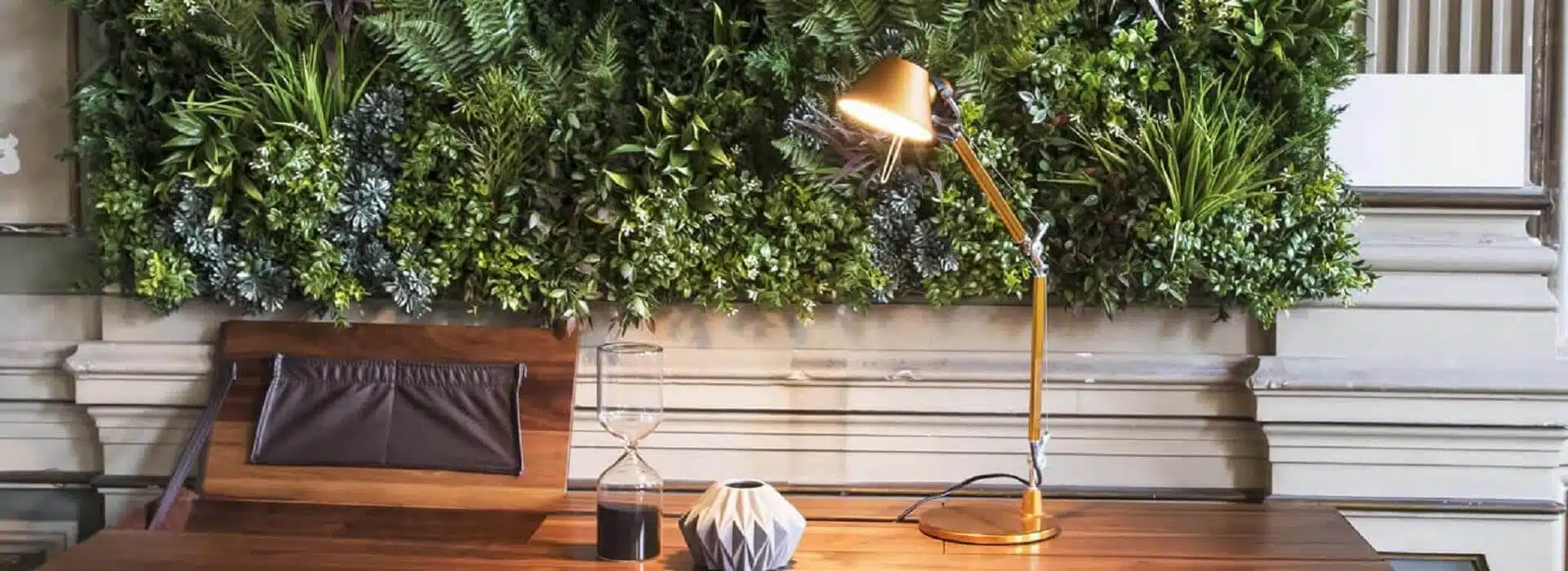 A wooden desk with a gold lamp, sand timer, and vase stands against a Home Green Wall Design in Lisbon, showcasing lush plants. The setting seamlessly blends modern and natural ambiance.