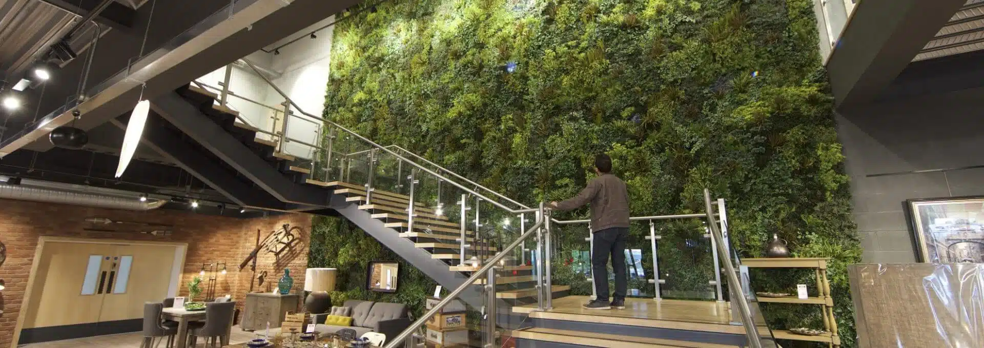 A person stands on a staircase inside a modern UK building, admiring a luxurious custom green wall in Yorkshire. The space blends industrial design elements with exposed brick and ductwork, while contemporary furniture is visible nearby.