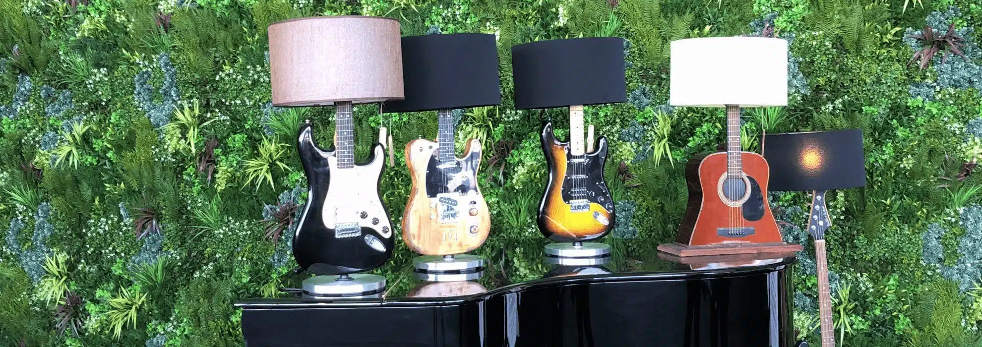 Four guitar lamps adorn a black piano against a lush green wall, capturing the essence of festival design. Three electric guitars boast varied finishes, while an acoustic guitar adds a sleek black shade. Perfect for any music event setup.