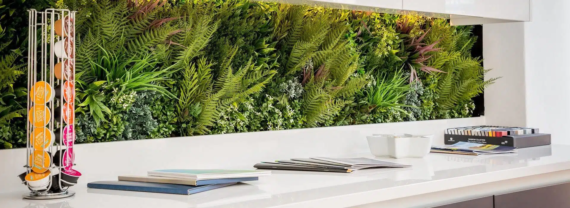 A modern office space with a white countertop displaying neatly stacked books and two cups. To the left, a stand holds colorful coffee pods. The backdrop is enhanced by stunning artificial green walls in Brighton, offering a lush vertical garden feel with various plants.