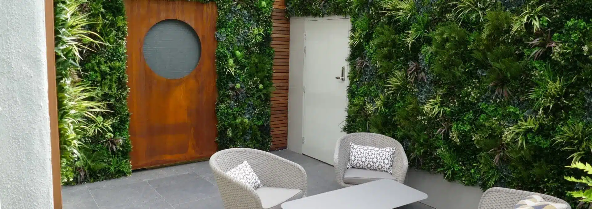 A modern outdoor patio with garden privacy screens in Fulham boasts a vertical garden on the walls. A round wooden door, two light gray wicker chairs with patterned cushions, and a matching table enhance this minimalist space surrounded by lush greenery.