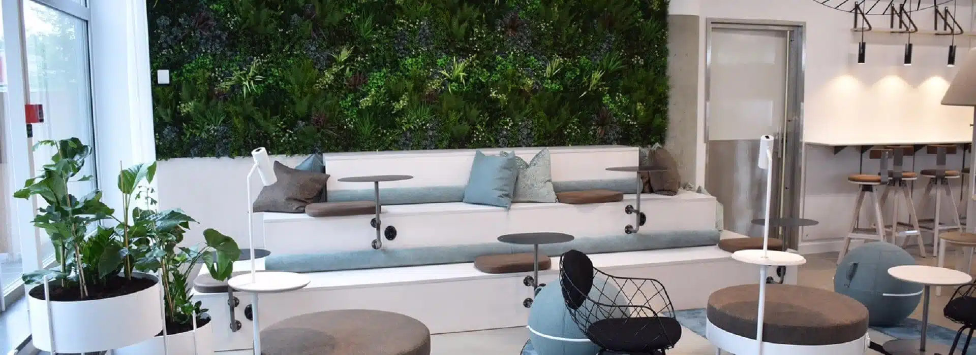Modern lounge area with tiered seating, small round tables, and wire chairs. A lush green wall reminiscent of trendy Swedish furniture stores adorns the space. Potted plants are scattered throughout, and bar stools line a counter in the back. The area is well-lit and contemporary.