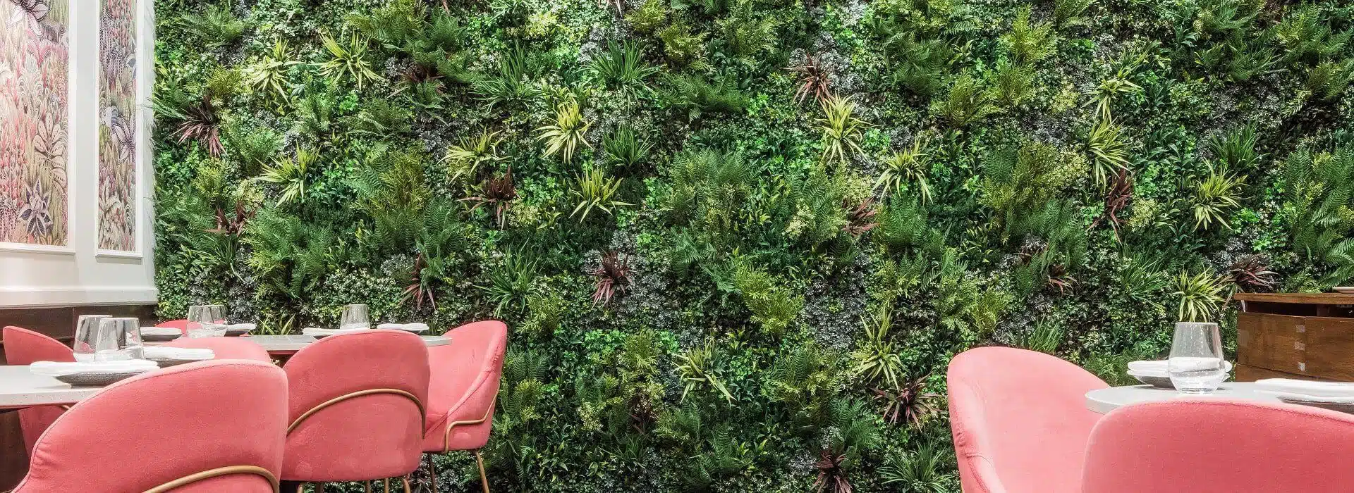 A London restaurant interior boasts an artificial green wall teeming with lush greenery. In the foreground, round tables adorned with glassware are surrounded by pink upholstered chairs, creating a vibrant and inviting dining atmosphere.