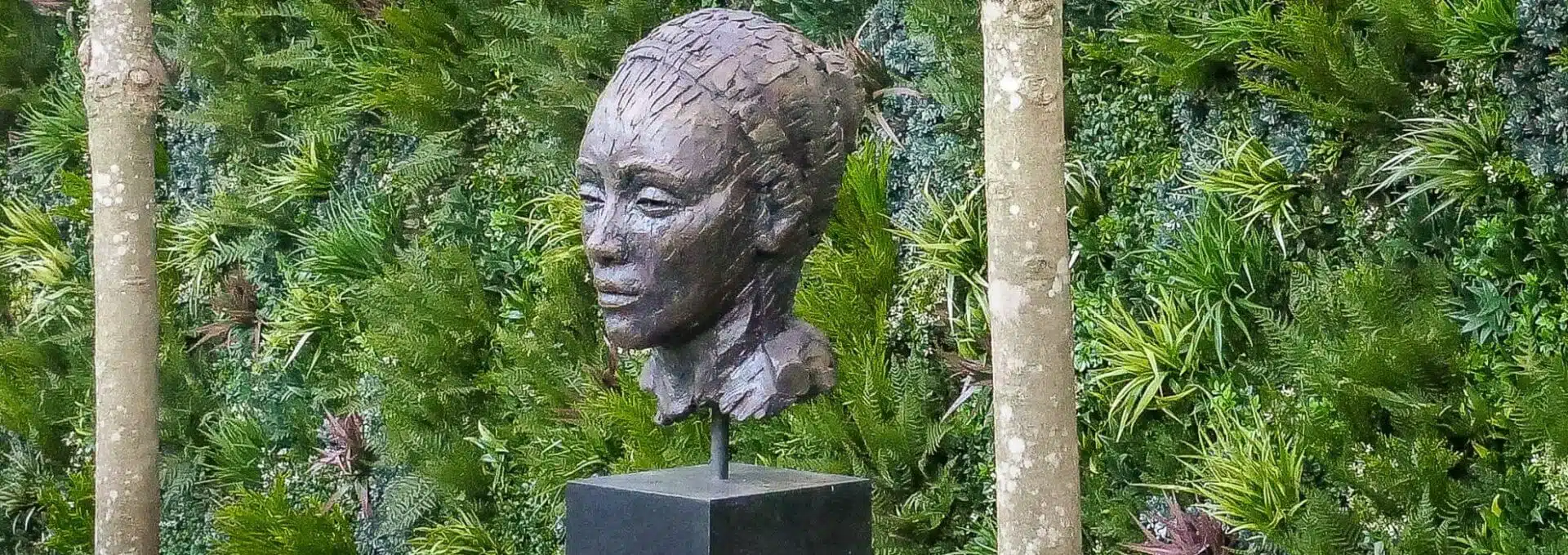 A luxurious sculpture garden features a bronze bust on a black pedestal, elegantly set against a lush green wall teeming with diverse plants, while two tree trunks gracefully flank the scene.
