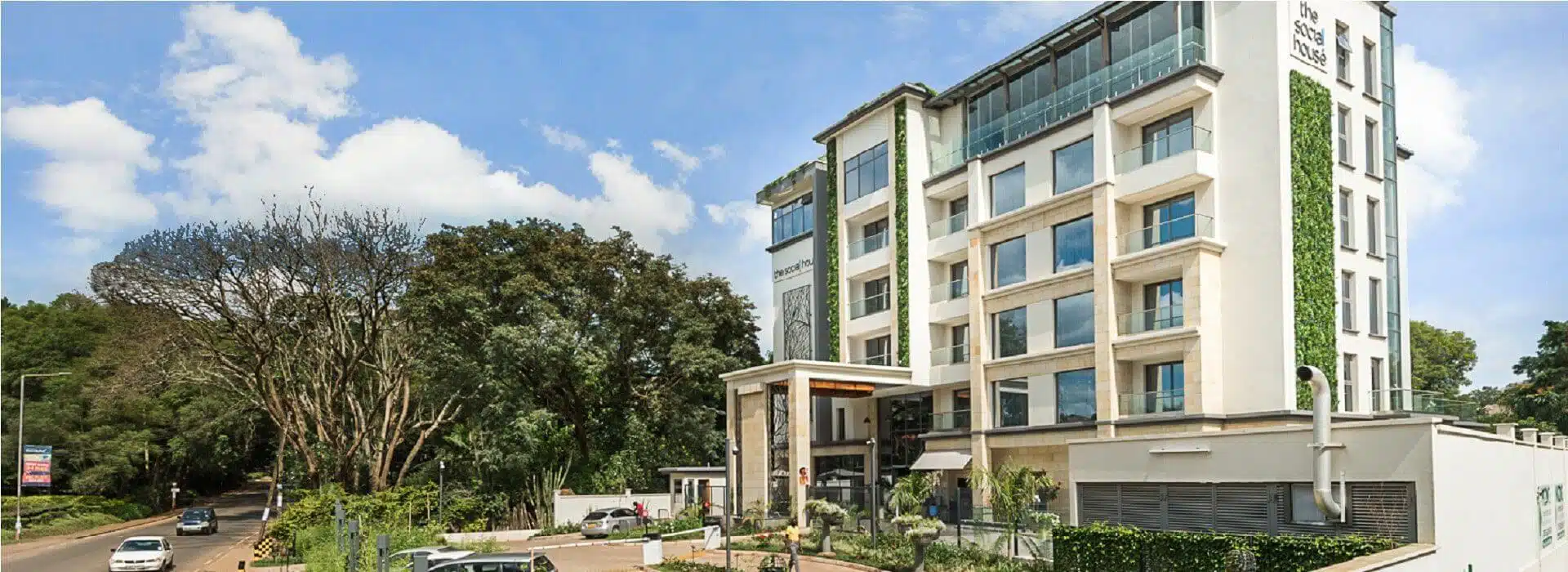 The Social House Hotel is a modern multi-story building in Nairobi, featuring large windows, green ivy accents, and a glass terrace on the top floor. Surrounded by lush trees and adjacent to a road with parked cars, it stands under a bright blue sky with scattered clouds.