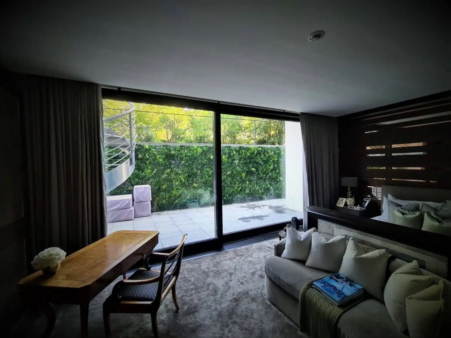 A modern living room with a large glass door opens to a patio, where the Hyde Park Green Wall Replacement enhances the view. Inside, a wooden desk with chairs, a plush sofa adorned with pillows, and soft lighting create an inviting space. A book rests casually on the sofa.