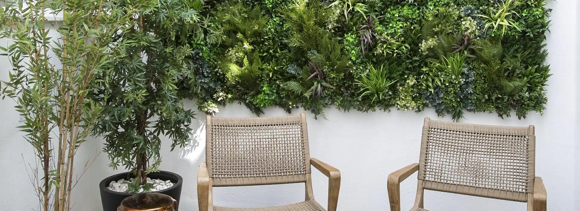 Two wooden chairs with woven backs are placed against a white wall adorned with a lush vertical garden. A potted plant with long, slender leaves is on the left, enhancing the UV-protected green wall privacy screen in the serene outdoor setting of Lisbon, Portugal.