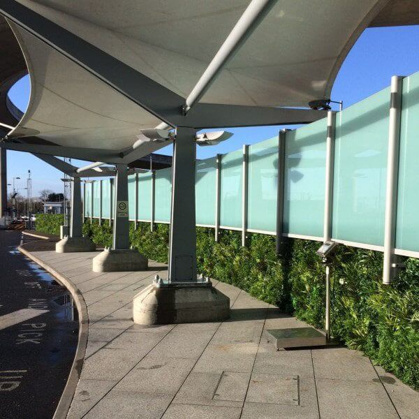 The exterior of Heathrow Airports Terminal 5 VIP Entrance features a modern, covered walkway. A fabric canopy supported by metal structures shades the path lined with glass panels and lush green walls, complemented by clear blue skies overhead.